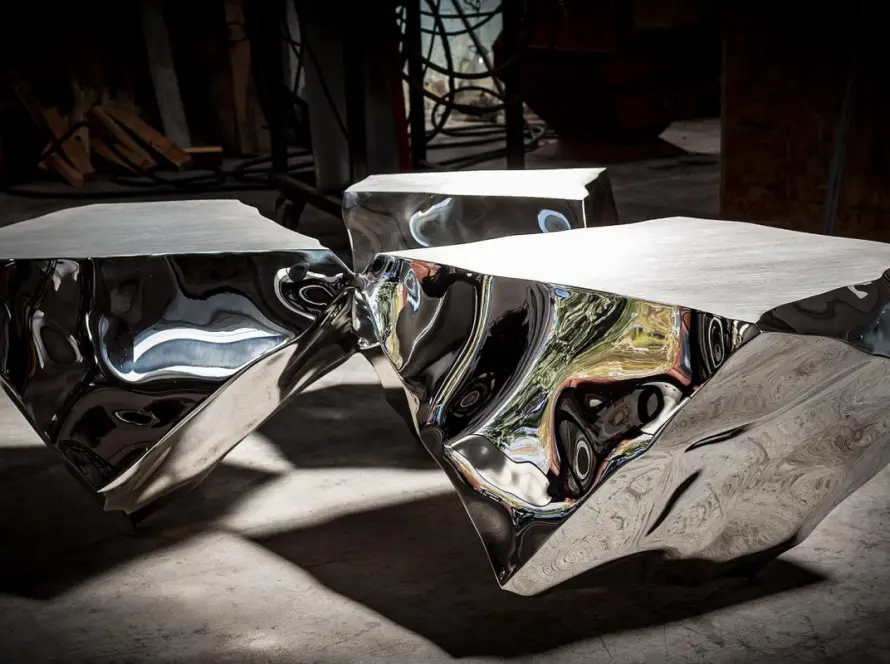 ALT: “The Connoisseurs” — three mirror-polished stainless-steel volumes at coffee-table height, derived from cube cross-sections, reflecting studio light; by Rado Kirov (2018). Caption: Three related volumes cut from a cube, where liquid reflections turn space into image.